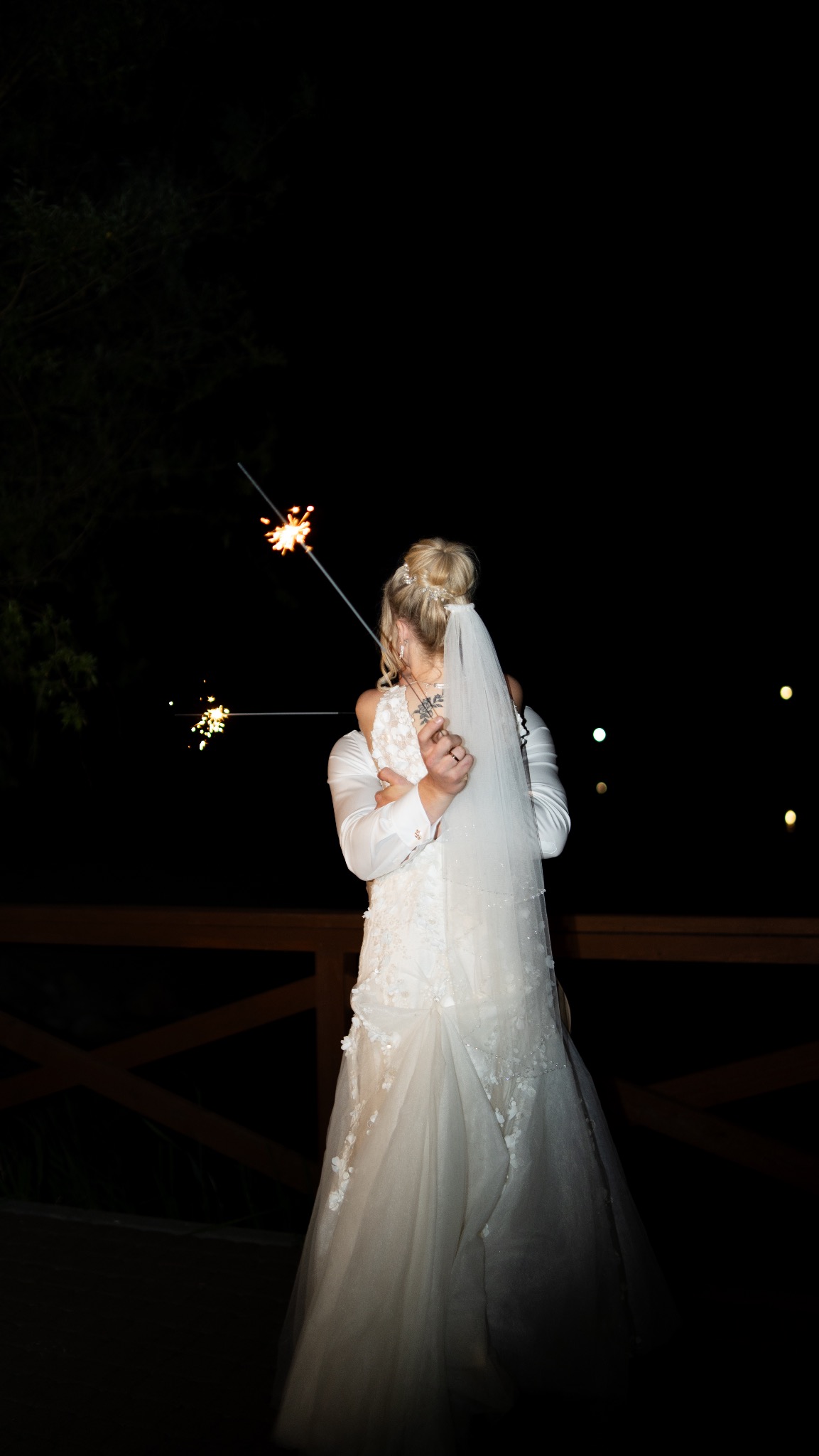 Para młoda obejmuje się na tle nocnego nieba, trzymając zapalone zimne ognie, widok z tyłu na koronkową suknię ślubną i welon.