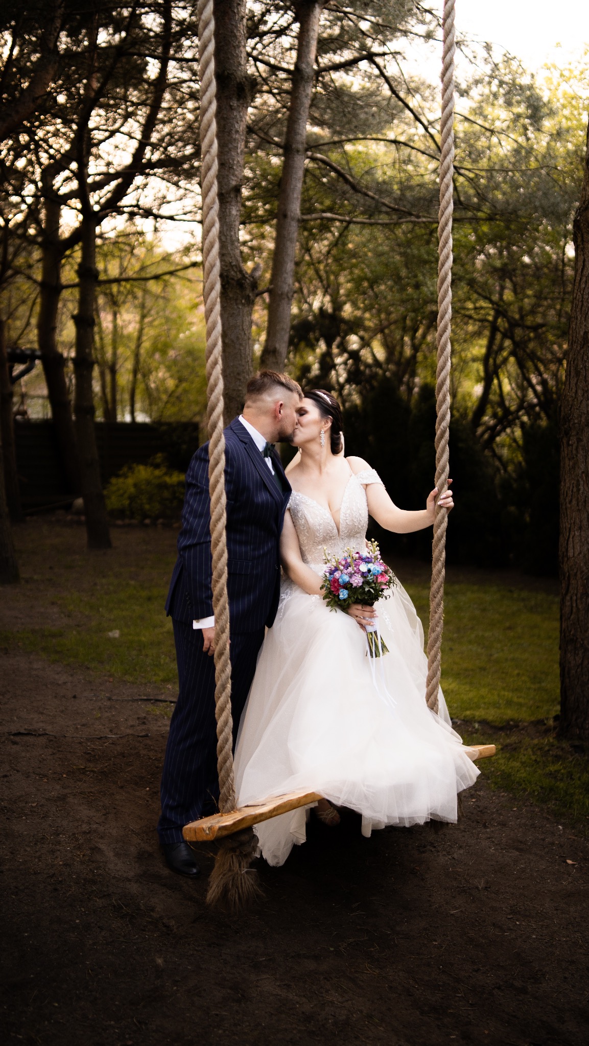 Panna młoda w białej sukni ślubnej i pan młody w granatowym garniturze w paski całują się na drewnianej huśtawce zawieszonej na linach w plenerze.