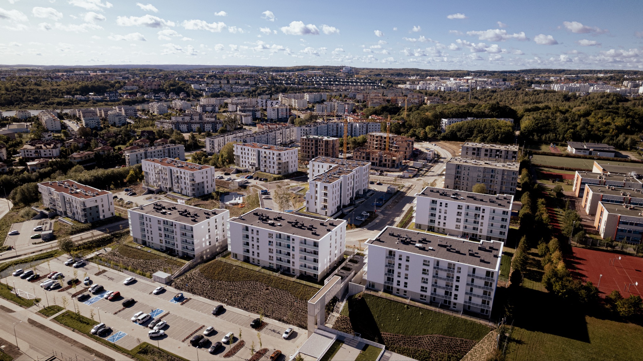 Widok z lotu ptaka na osiedle nowoczesnych, białych budynków mieszkalnych z balkonami, z parkingiem pełnym samochodów, otoczone zielenią i zabudową miejską w słoneczny dzień.