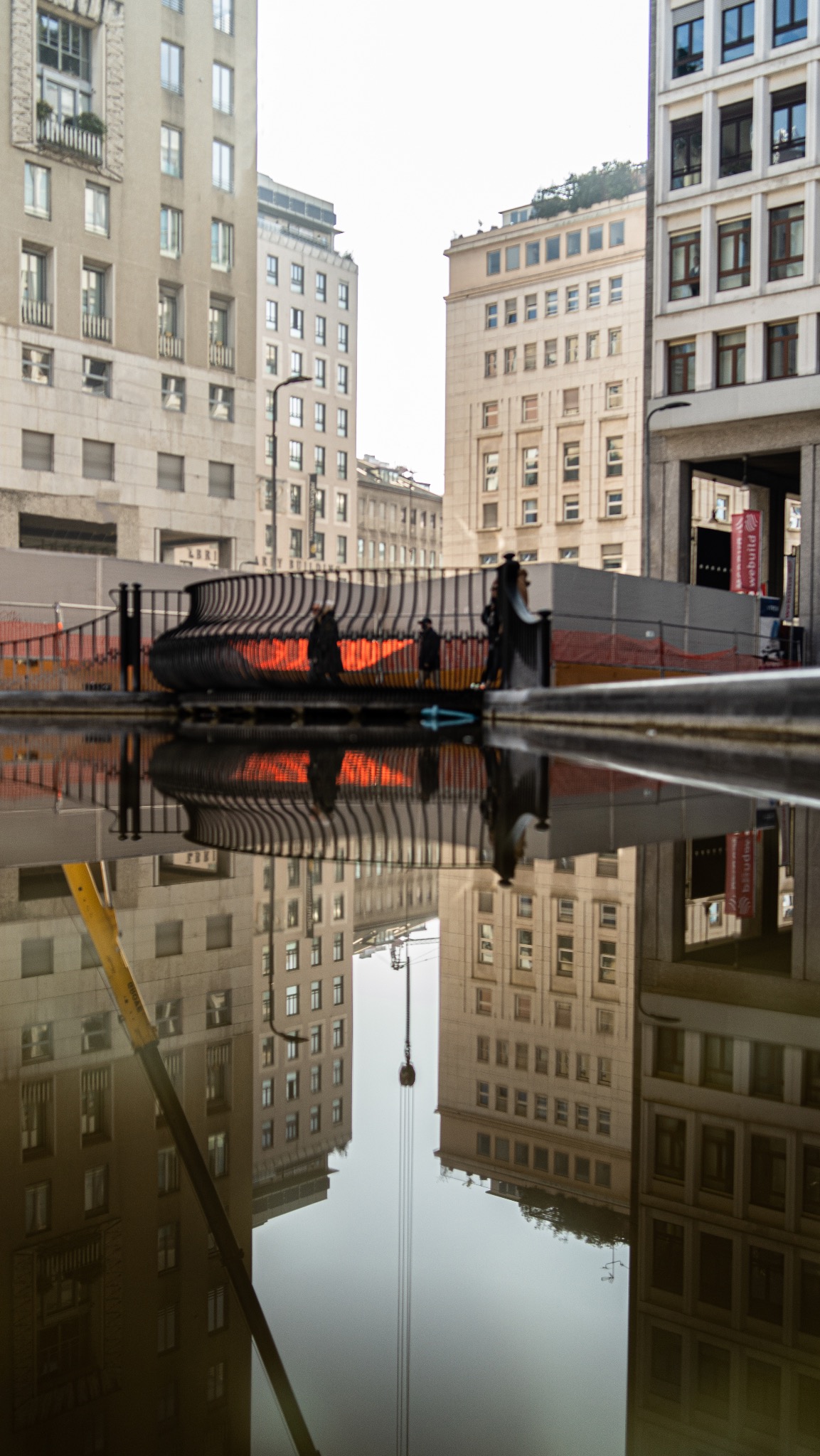 Architektura miejska odbita w kałuży: budynki, ażurowa konstrukcja i sylwetki przechodniów.