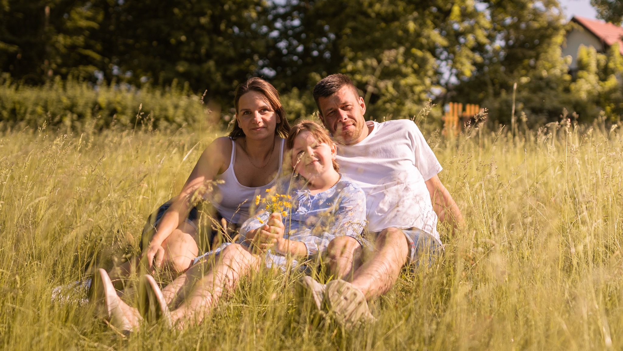 Rodzinne zdjęcie: mama, tata i córka siedzą na łące w wysokiej trawie, dziewczynka trzyma bukiecik żółtych kwiatów, w tle drzewa i dom z czerwonym dachem.