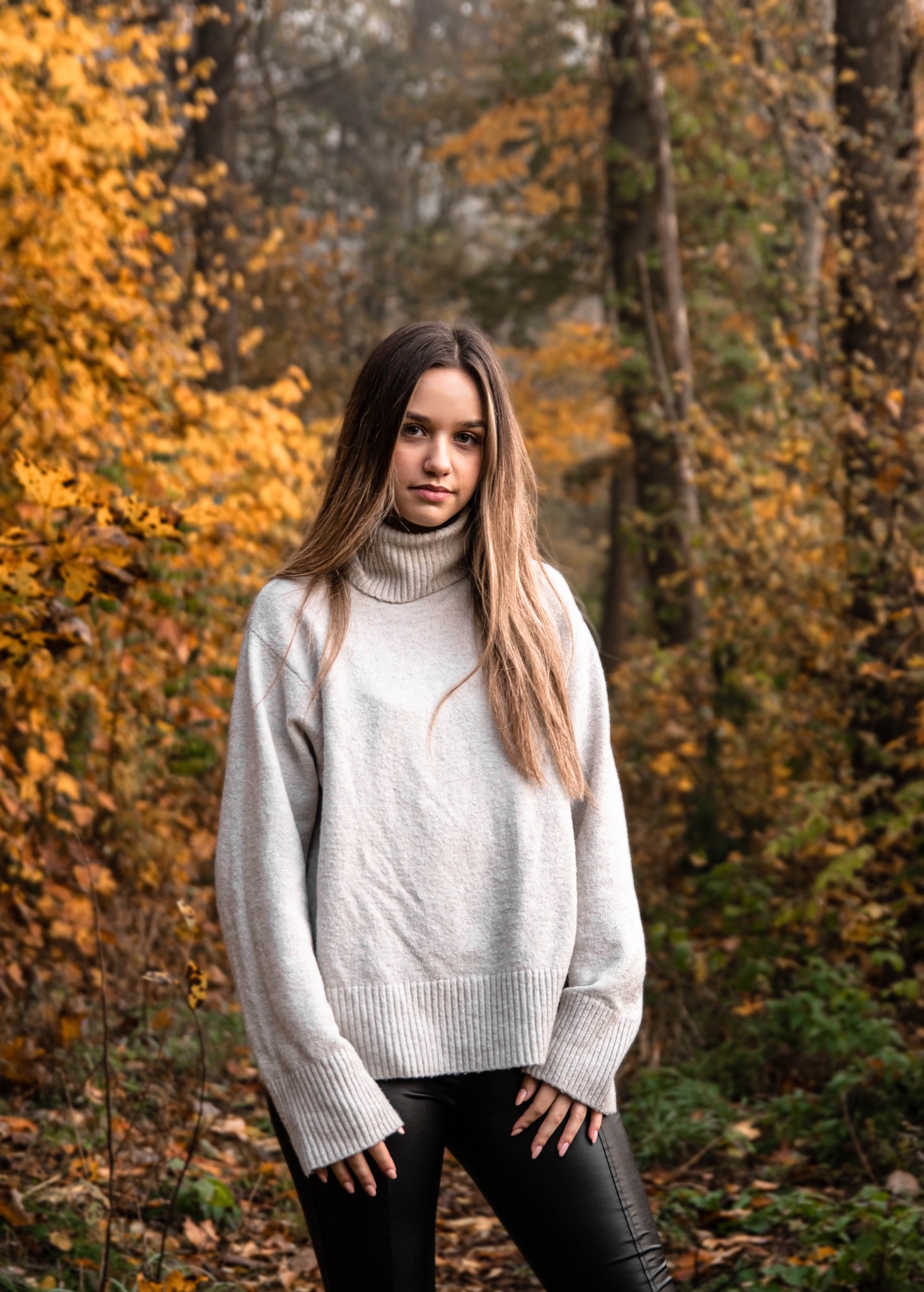 Portret młodej kobiety w swetrze z golfem i skórzanych spodniach, na tle jesiennego lasu z żółtymi liśćmi.