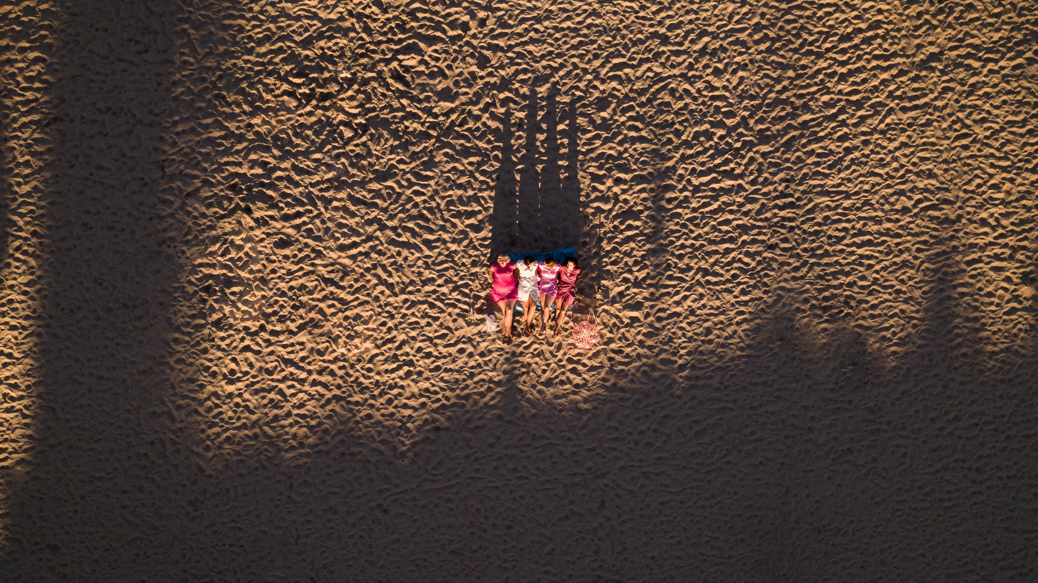 Widok z góry na cztery kobiety leżące na kocu na piaszczystej plaży, rzucające długie cienie w popołudniowym słońcu.