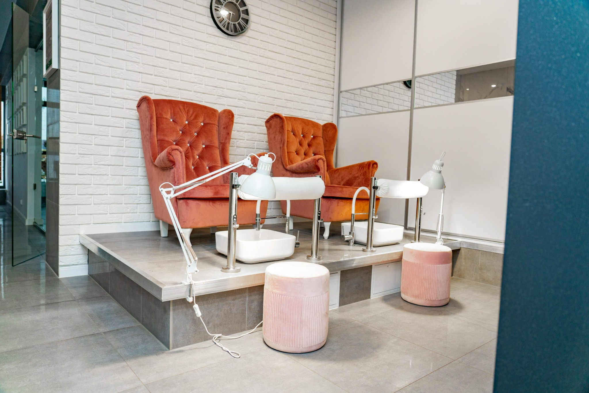 Dwa fotele w kolorze terakoty z pikowaniem, stanowiska do pedicure z białymi umywalkami i lampami, różowe pufy na tle białej ceglanej ściany i szafy z lustrzanymi wstawkami.