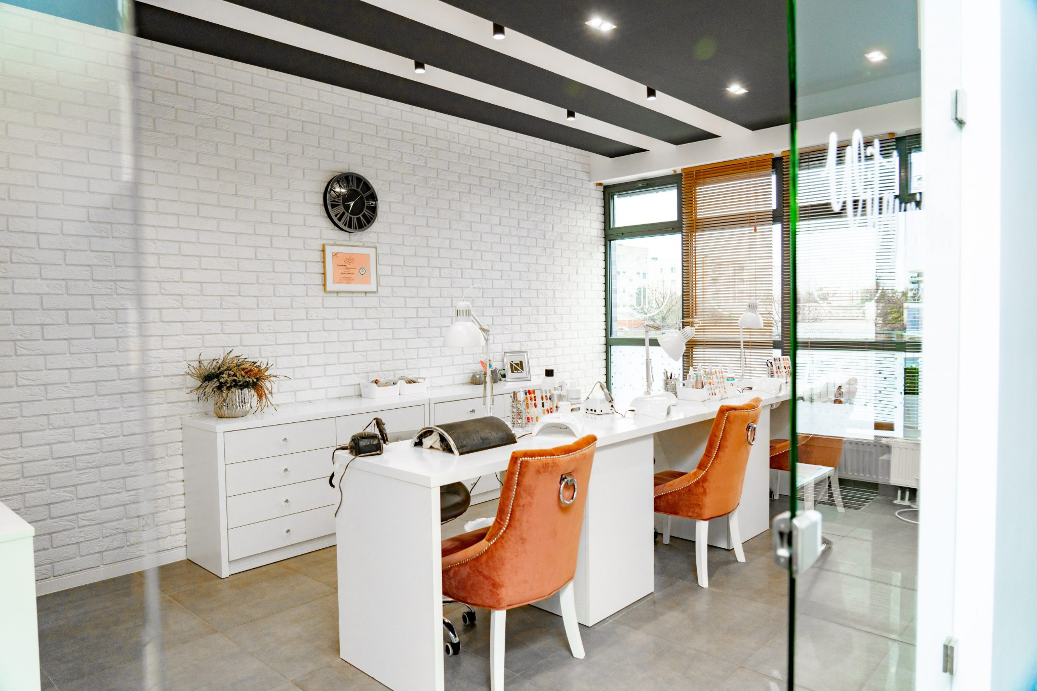 Wnętrze salonu kosmetycznego: białe stanowiska do manicure z pomarańczowymi krzesłami, biała ceglana ściana z zegarem i dekoracją, szara podłoga, czarny sufit z oświetleniem punktowym.