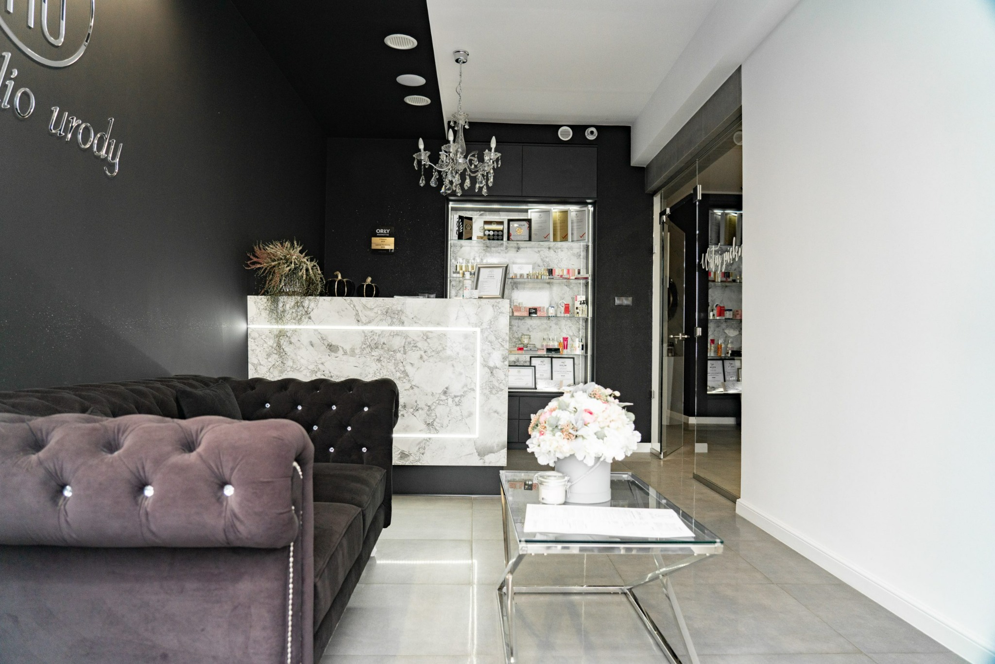 Eleganckie wnętrze salonu urody z marmurową recepcją podświetloną LED, kryształowym żyrandolem i aksamitną sofą w odcieniach szarości, uzupełnione dekoracjami i szklanym stolikiem.