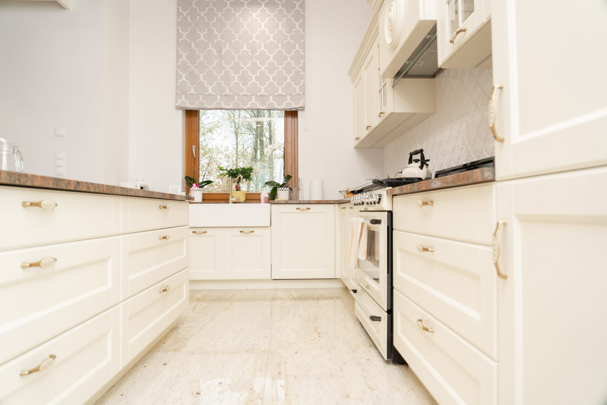 Jasna kuchnia w stylu klasycznym z beżowymi szafkami, granitowym blatem i oknem z roletą rzymską w geometryczny wzór.