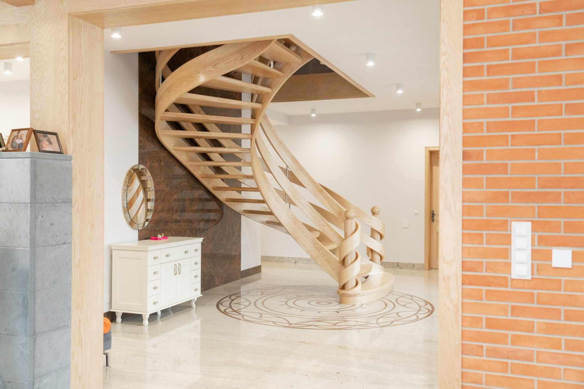 Wnętrze domu w Koninie z drewnianymi, spiralnymi schodami, marmurową posadzką z dekoracyjnym wzorem, białą komodą i ceglaną ścianą w tle.