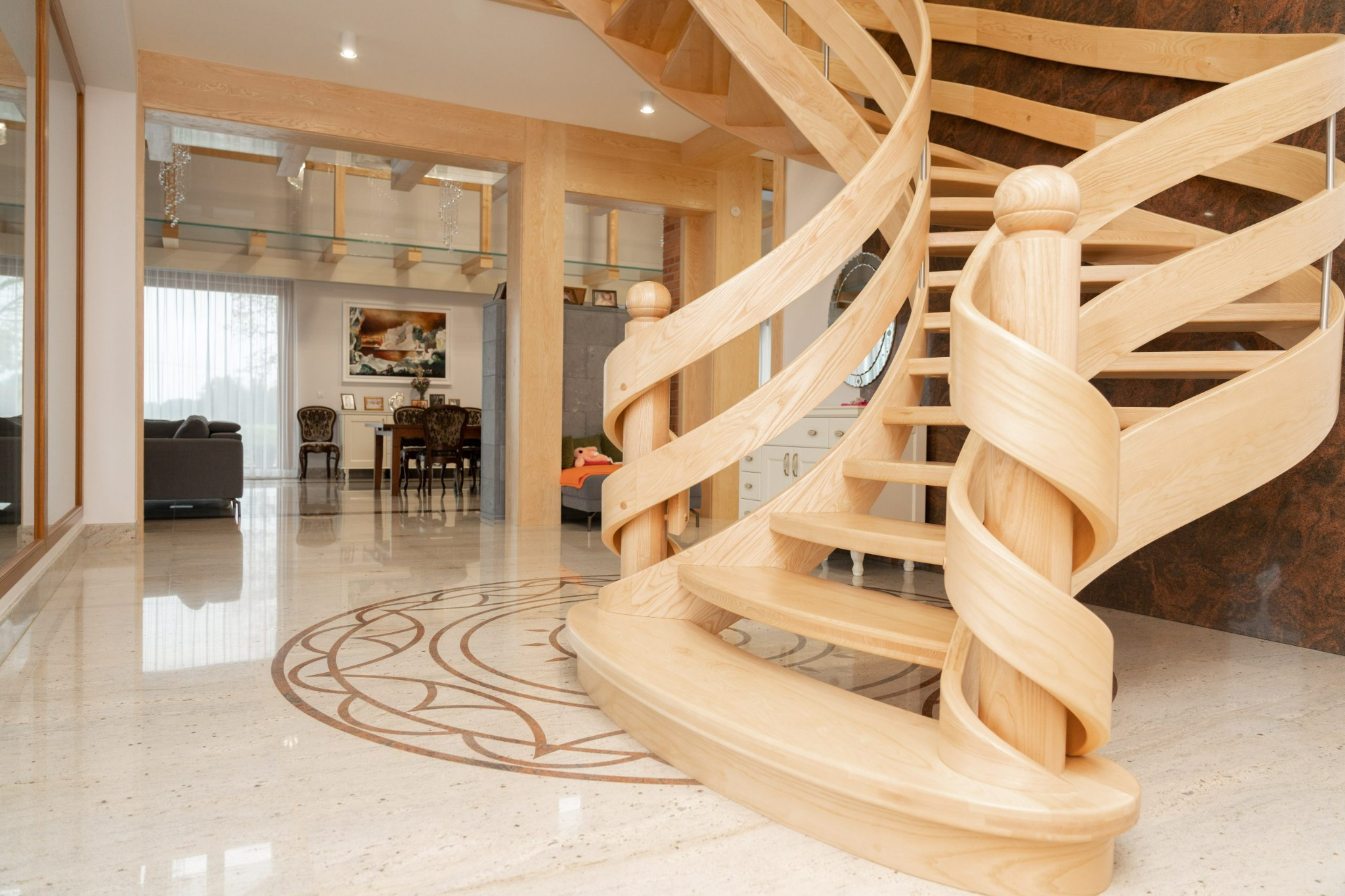 Spiralne, drewniane schody z jasnego drewna w przestronnym holu z marmurową podłogą i dekoracyjnym wzorem. W tle widoczne meble i elementy wystroju wnętrz.