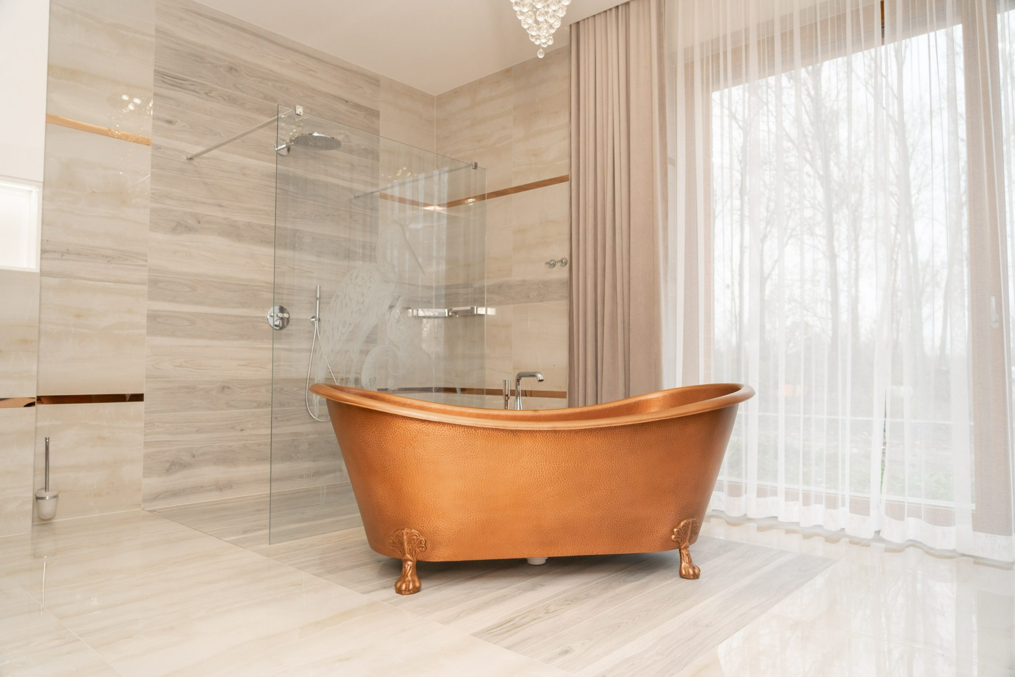 Luksusowa łazienka z miedzianą wanną na nóżkach, szklaną kabiną prysznicową i marmurowymi płytkami w odcieniach beżu, oświetlona kryształowym żyrandolem i naturalnym światłem z okna z delikatnymi...