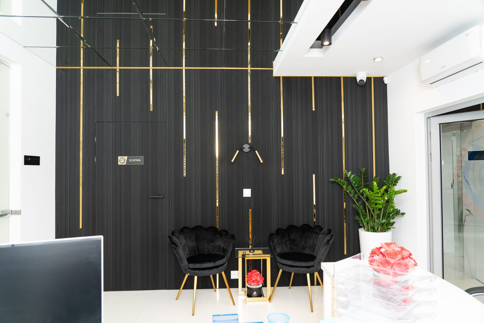 Recepcja biura z czarną ścianą zdobioną złotymi listwami, czarnymi fotelami muszelkami na złotych nogach i dekoracjami kwiatowymi.