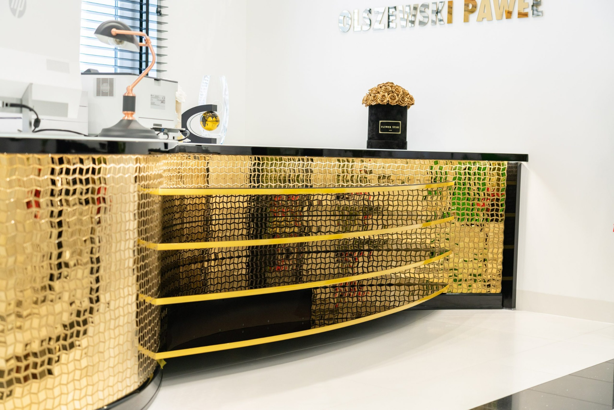 Recepcja biurowa z dekoracyjną, złotą mozaiką na froncie, czarnym blatem i elementami dekoracyjnymi: lampką, globusem i bukietem róż w czarnym pudełku.