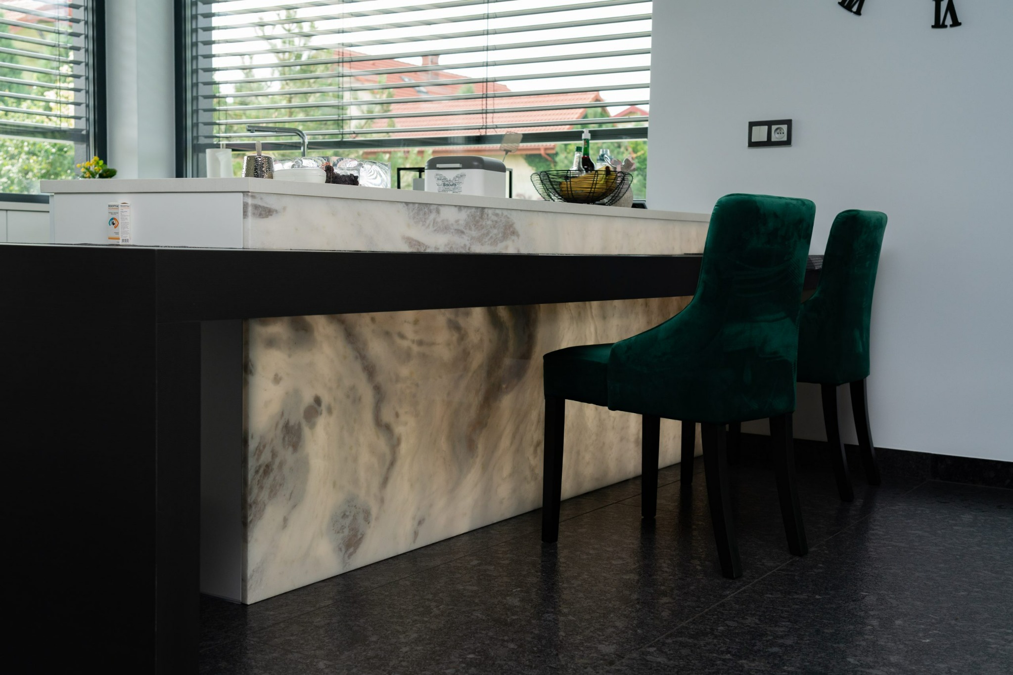 Wyspa kuchenna z podświetlanym, marmurkowym frontem i dwoma ciemnozielonymi, welurowymi krzesłami barowymi; widok na okno z żaluzjami i elementy dekoracyjne na blacie.