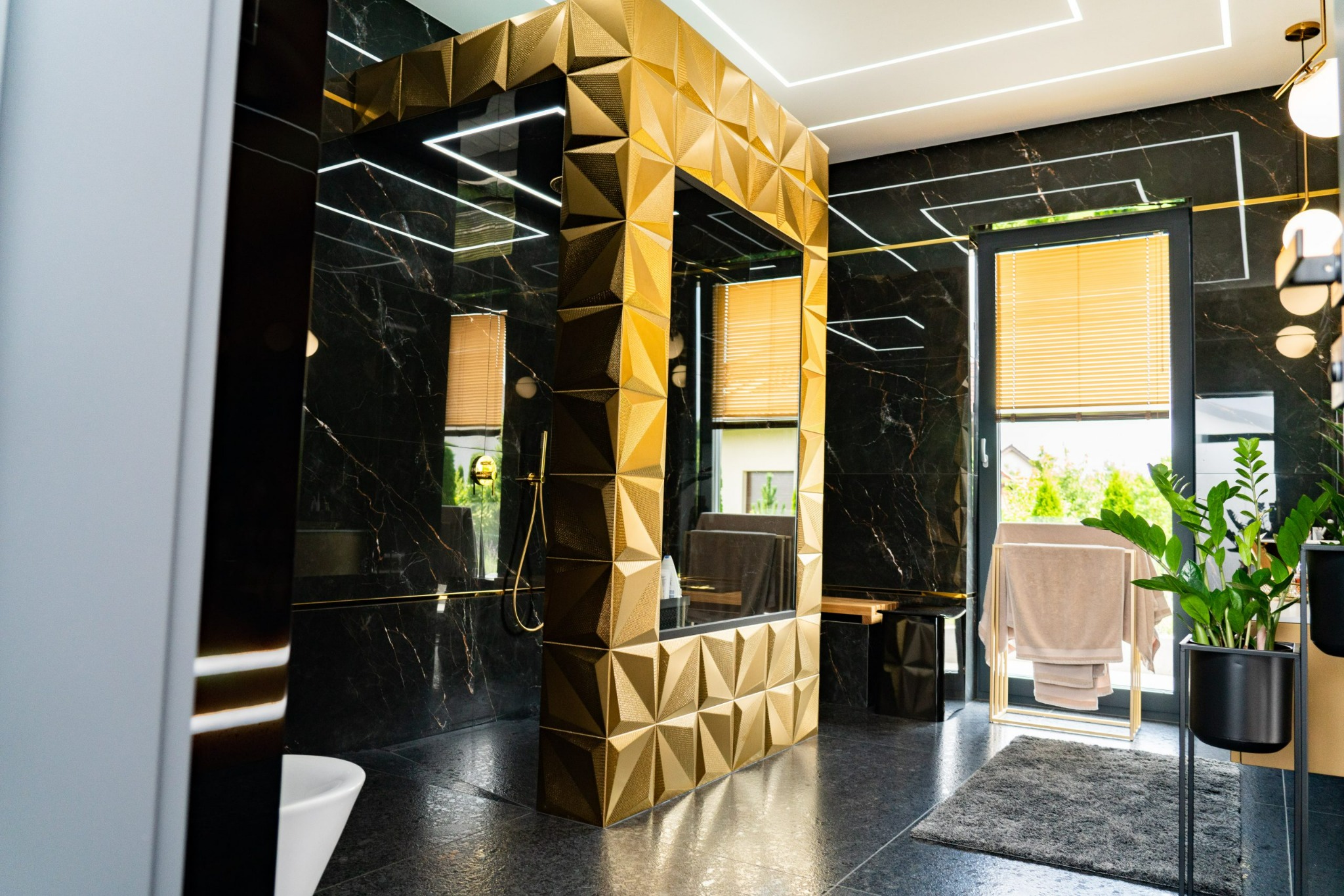 Luksusowa łazienka z czarnymi marmurowymi ścianami, złotą kabiną prysznicową o geometrycznym wzorze, oświetleniem LED i roślinami doniczkowymi.