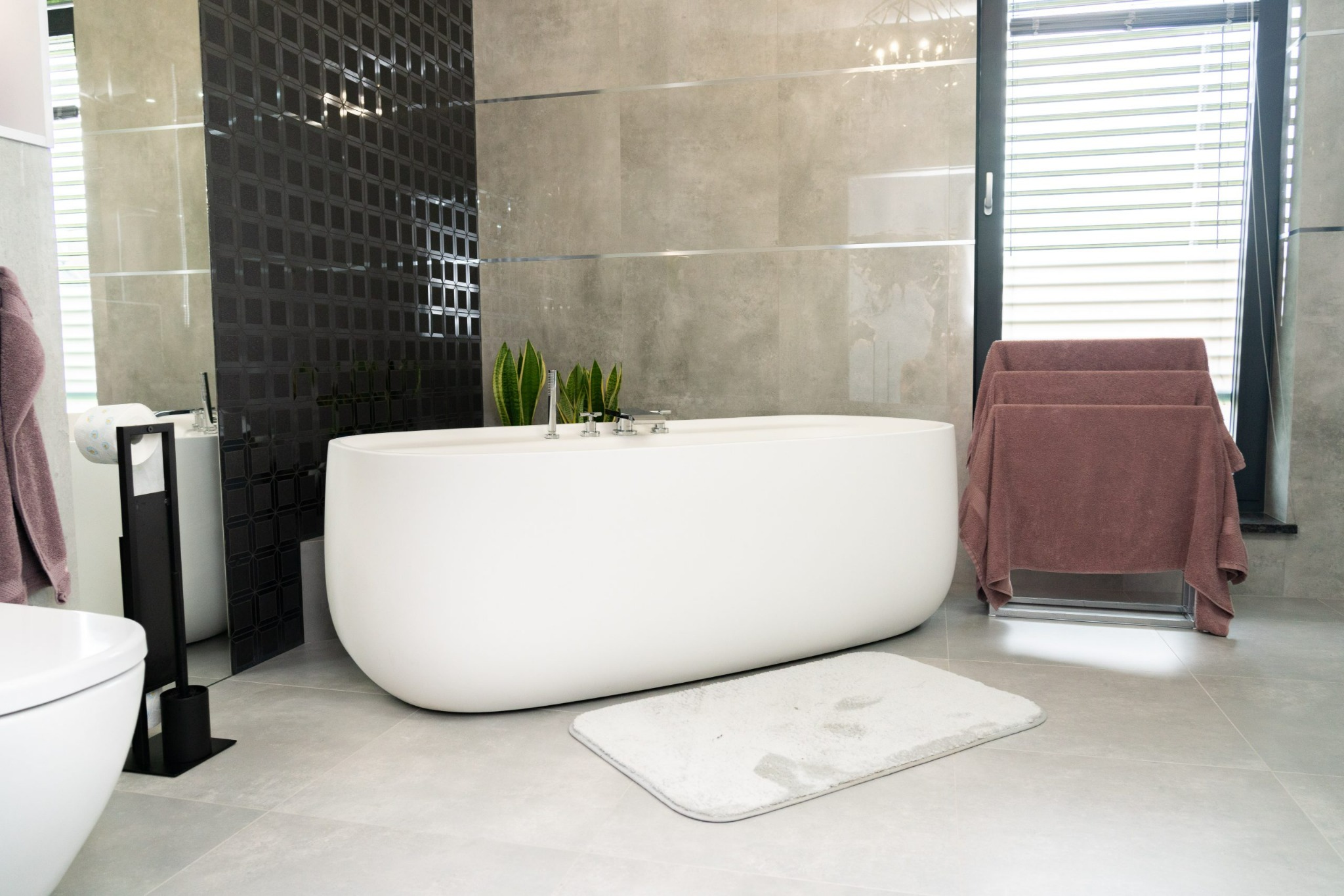 Nowoczesna łazienka z wolnostojącą białą wanną, szarą płytką, czarną mozaiką i roślinami w tle.