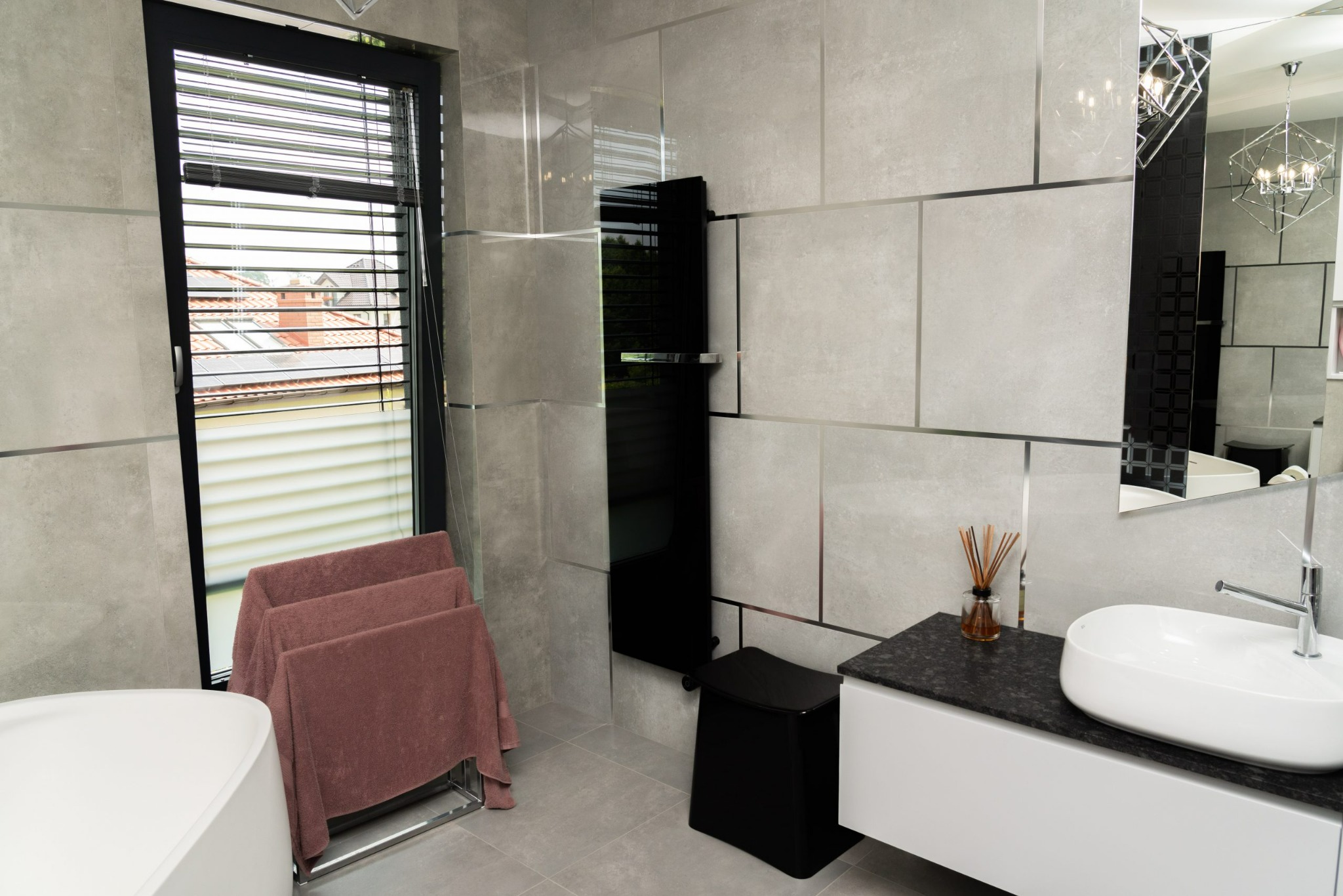 Nowoczesna łazienka z szarymi płytkami, czarnym grzejnikiem drabinkowym i owalną umywalką nablatową na białej szafce z czarnym blatem. W tle widoczne okno z żaluzjami i dekoracyjna lampa.