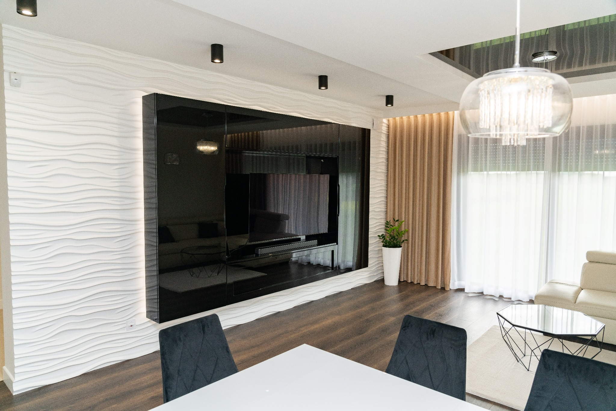 Nowoczesny salon z czarną ścianką RTV, trójwymiarową białą ścianą, szklanym żyrandolem i drewnianą podłogą, widziany z perspektywy stołu z szarymi krzesłami.