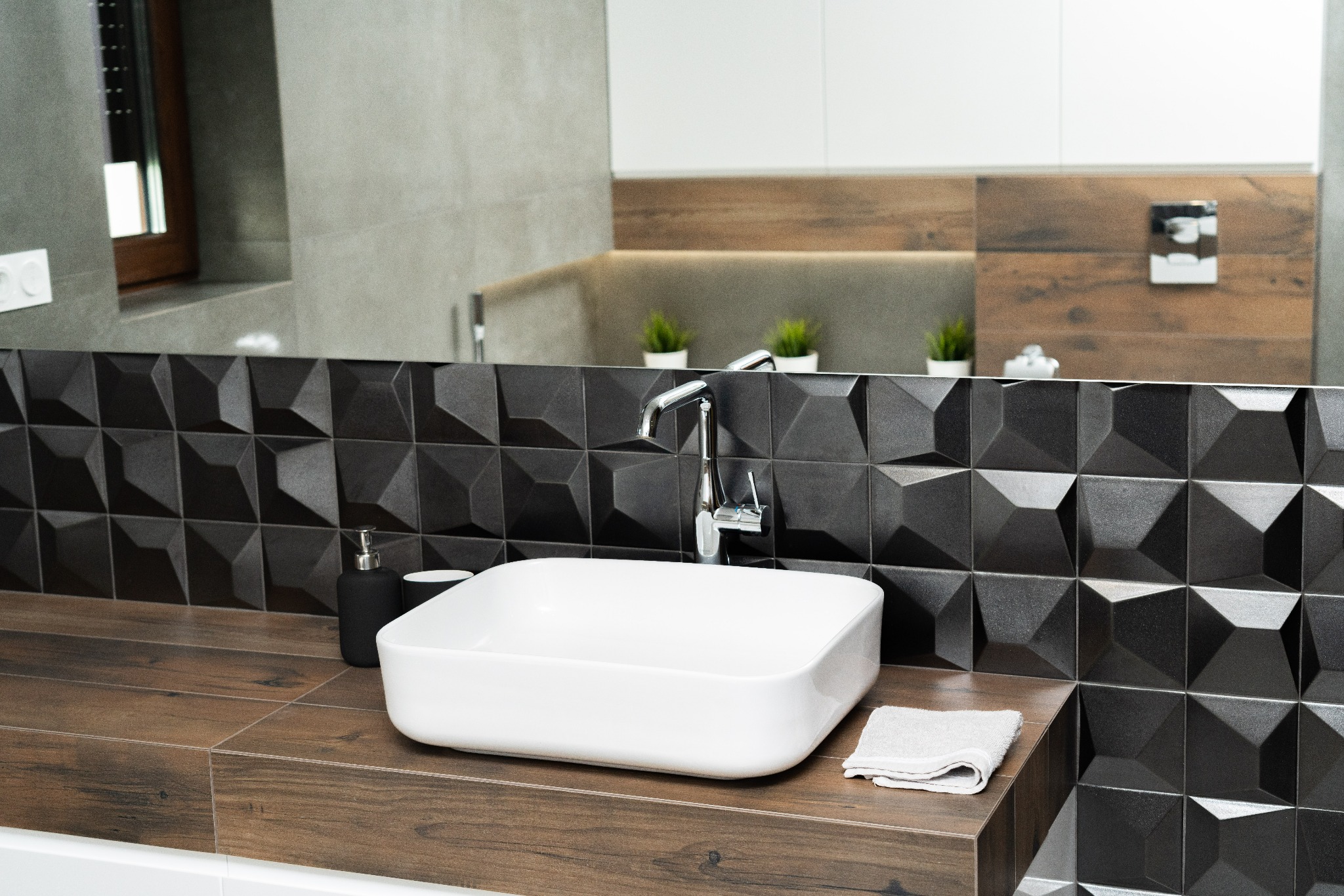 Nowoczesna łazienka z białą, kwadratową umywalką nablatową, chromowaną baterią i czarną, trójwymiarową mozaiką na ścianie. Blat z płytek imitujących drewno. W tle widoczne rośliny doniczkowe.