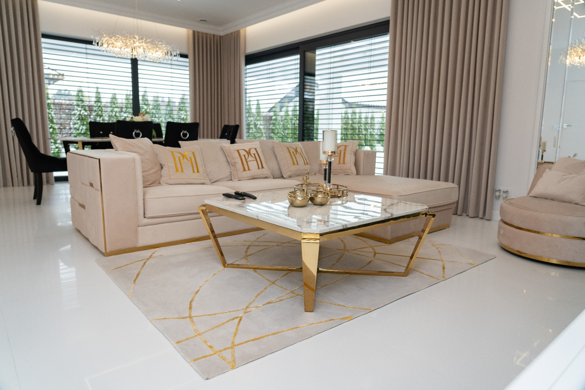 Jasny salon z beżową sofą z poduszkami z monogramem, marmurowym stolikiem kawowym ze złotymi nogami i dywanem w geometryczne wzory, widok na jadalnię i okno z żaluzjami.