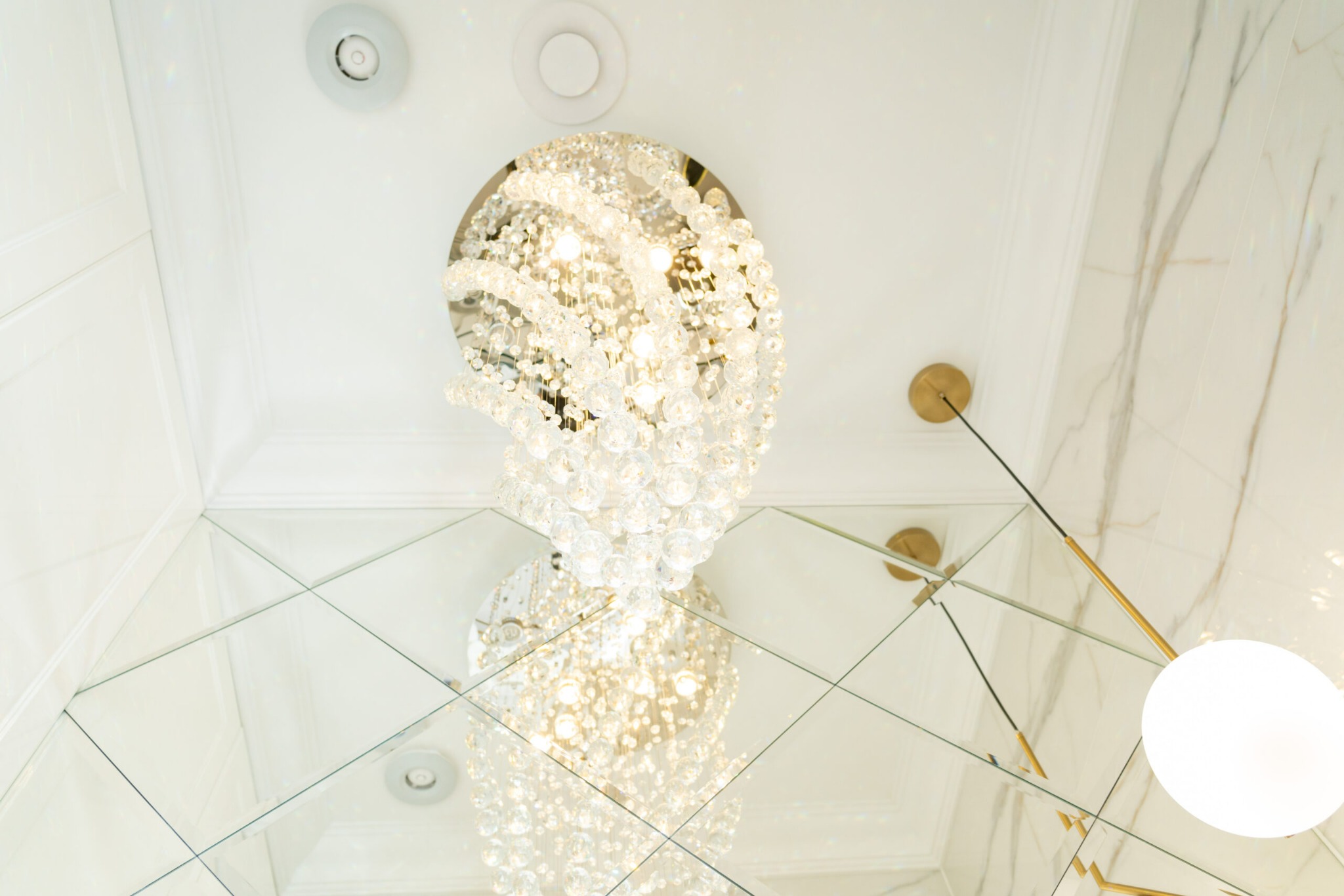 Kryształowy żyrandol w kształcie kuli, odbijający się w lustrzanym suficie z geometrycznym wzorem, oświetlenie punktowe i dekoracyjne elementy w kolorze złota.