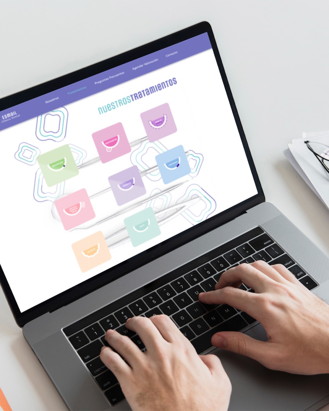 Osoba pisze na laptopie, na ekranie widoczna strona internetowa z kolorowymi ikonami i napisem NUESTROS TRATAMIENTOS, obok laptopa leżą okulary i dokumenty.
