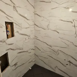 Fragment łazienki w trakcie prac wykończeniowych, widoczne białe płytki imitujące marmur na ścianach, ciemna podłoga, niezamaskowane otwory na instalacje oraz licznik wody w ścianie.