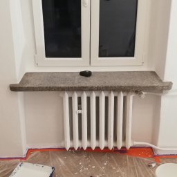 Paweł Rzeszot - Biały grzejnik pod oknem z kamiennym parapetem, zabezpieczona folią podłoga z narzędziami malarskimi i puszką farby, świeżo pomalowane ściany.