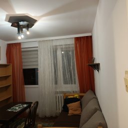 Paweł Rzeszot - Przytulne wnętrze pokoju z drewnianym regałem, biurkiem i sofą, okno z zasłonami i designerska lampa sufitowa. Widok na ścianę z włącznikiem światła.