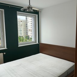 Paweł Rzeszot - Sypialnia po malowaniu we Wrocławiu: jedna ściana biała, druga butelkowa zieleń. Widok z okna na blok mieszkalny. Nowoczesna lampa sufitowa.