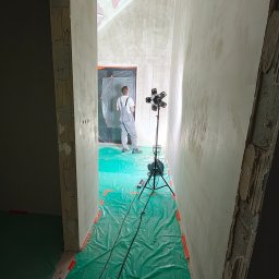 Paweł Rzeszot - Prace wykończeniowe we Wrocławiu: szpachlowanie ścian w korytarzu, zabezpieczona podłoga folią, widoczny pracownik i lampa oświetleniowa na statywie.