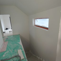 Paweł Rzeszot - Wnętrze domu w trakcie wykańczania: gładkie, białe ściany, zabezpieczone okno, schody w surowym stanie i szlifierka do gładzi na podłodze.