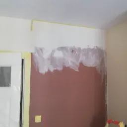 Pokój w trakcie prac przygotowawczych do malowania: zabezpieczone folią drzwi, ściana częściowo pokryta białą farbą gruntującą na brązowym podłożu, widoczne ślady szpachlowania.