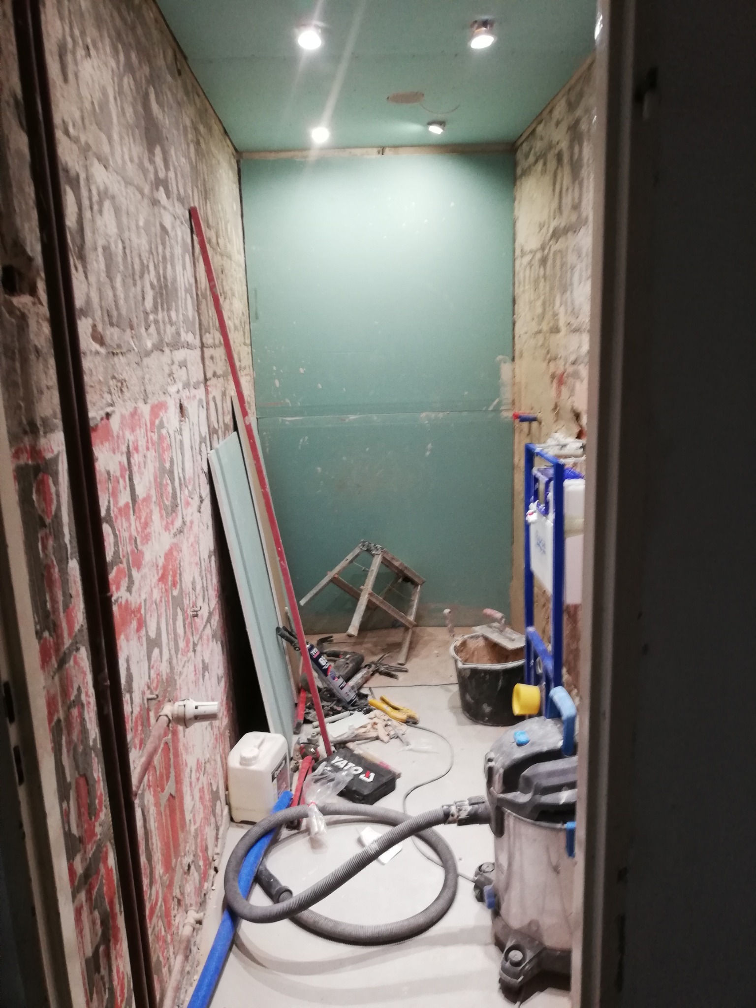 Łazienka w trakcie remontu: surowe ściany z pozostałościami farby, częściowo pokryte płytami gipsowo-kartonowymi, widoczny stelaż podtynkowy WC, narzędzia i odkurzacz przemysłowy na podłodze.