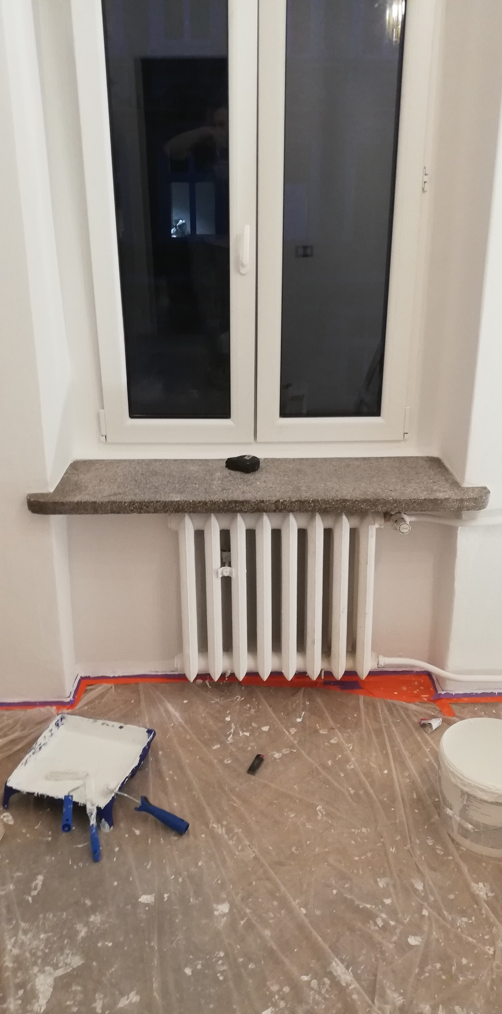 Biały grzejnik pod oknem z kamiennym parapetem, zabezpieczona folią podłoga z narzędziami malarskimi i puszką farby, świeżo pomalowane ściany.