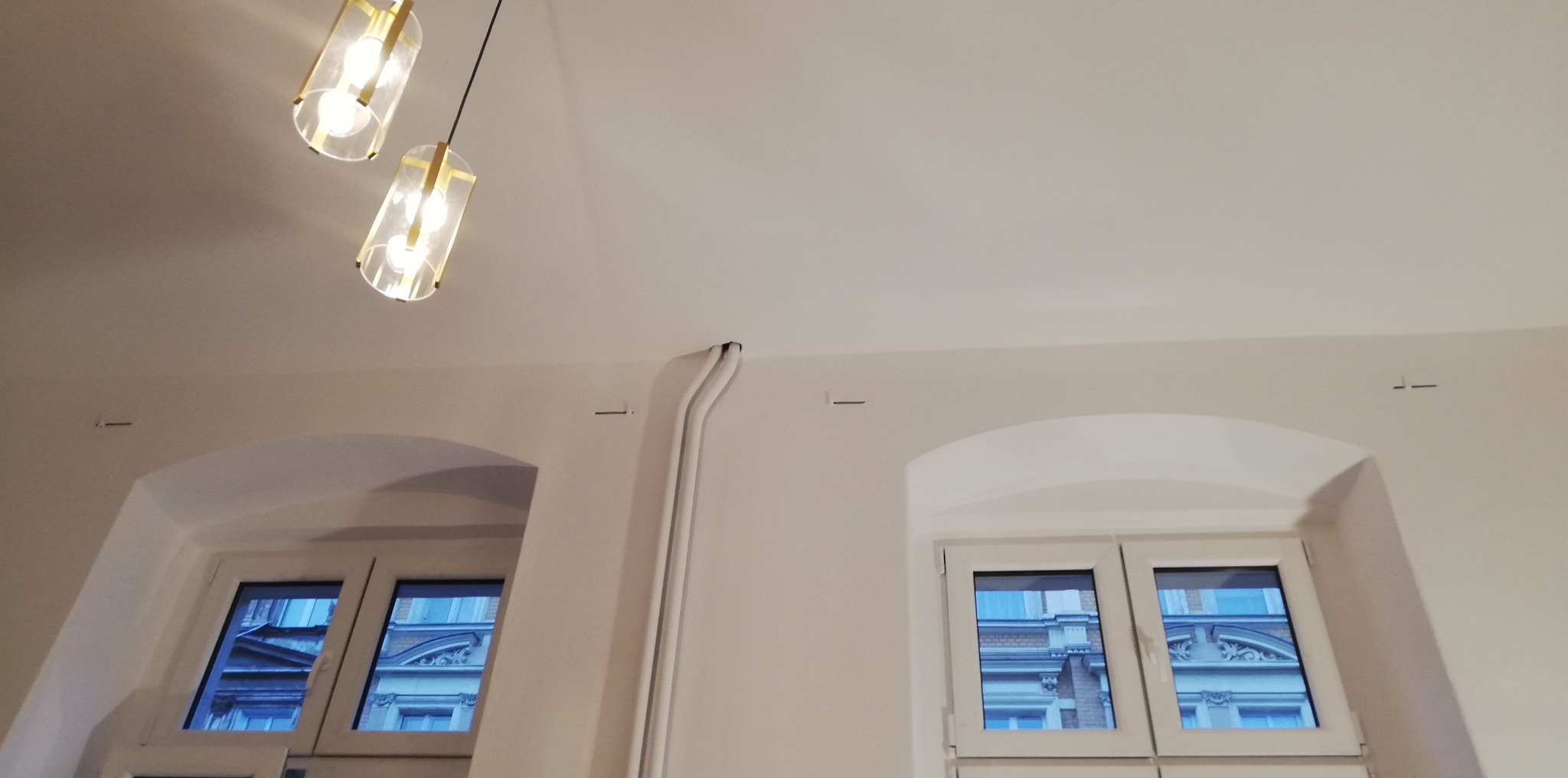 Świeżo pomalowane wnętrze z dwoma oknami w łukowych wnękach, widoczne kable w białej listwie i nowoczesne lampy wiszące z złotymi akcentami.
