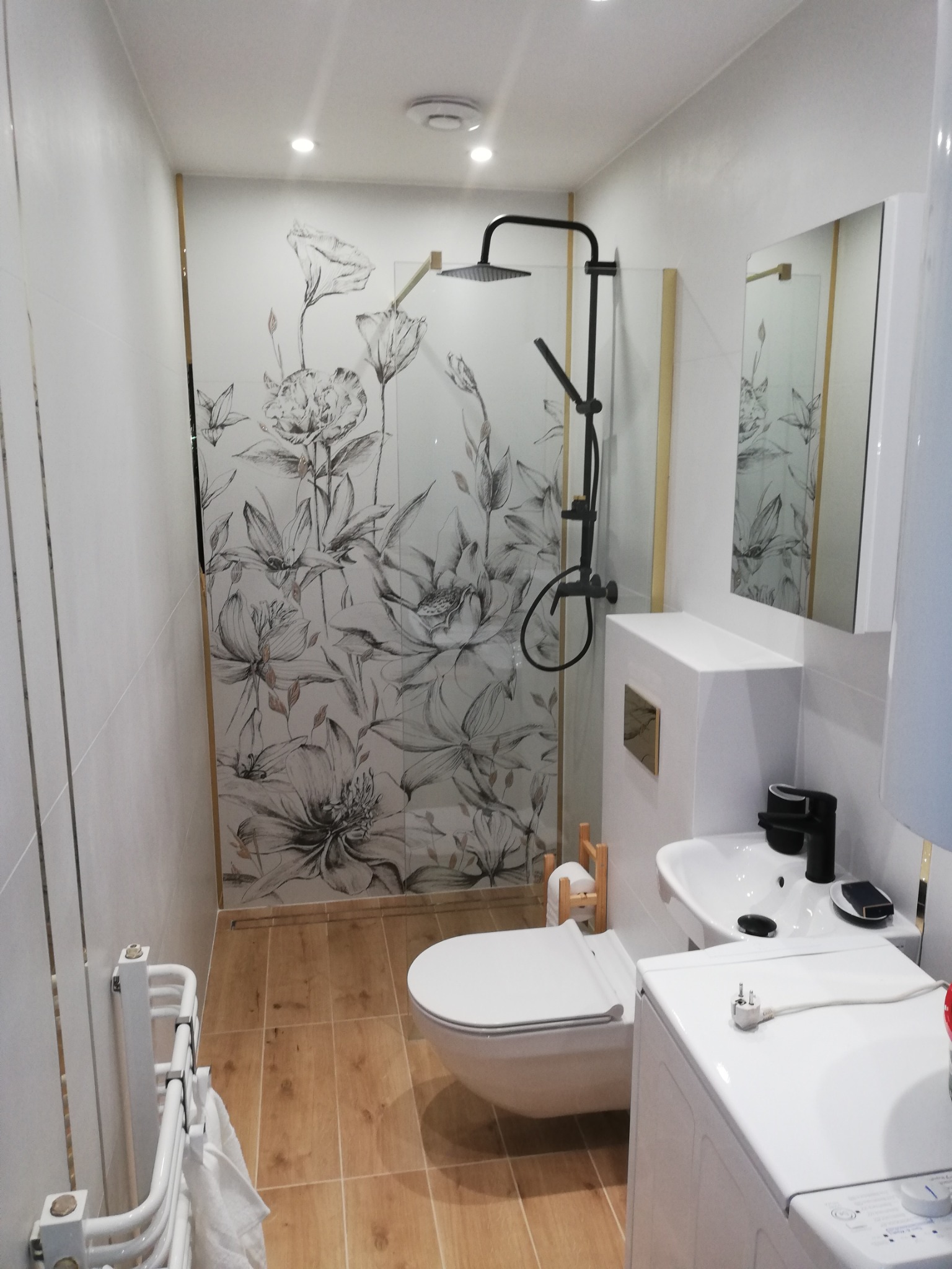 Nowoczesna łazienka po remoncie z kabiną prysznicową z motywem kwiatowym, czarną armaturą i drewnopodobnymi płytkami na podłodze.