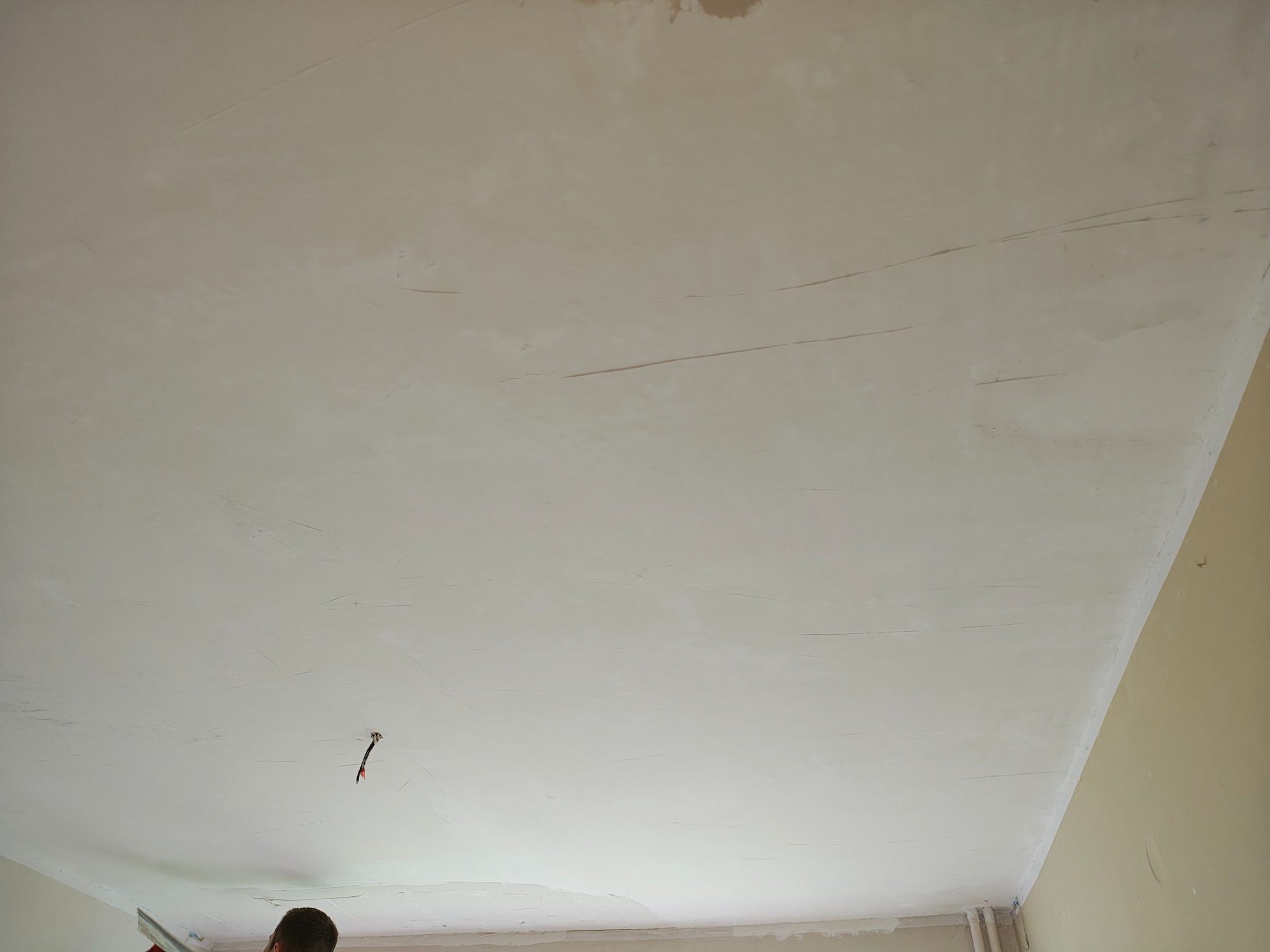 Niedokończone szpachlowanie sufitu z widocznymi smugami i fragmentem osoby w dolnym kadrze, uchwycone z perspektywy żabiej z widocznym kablem.