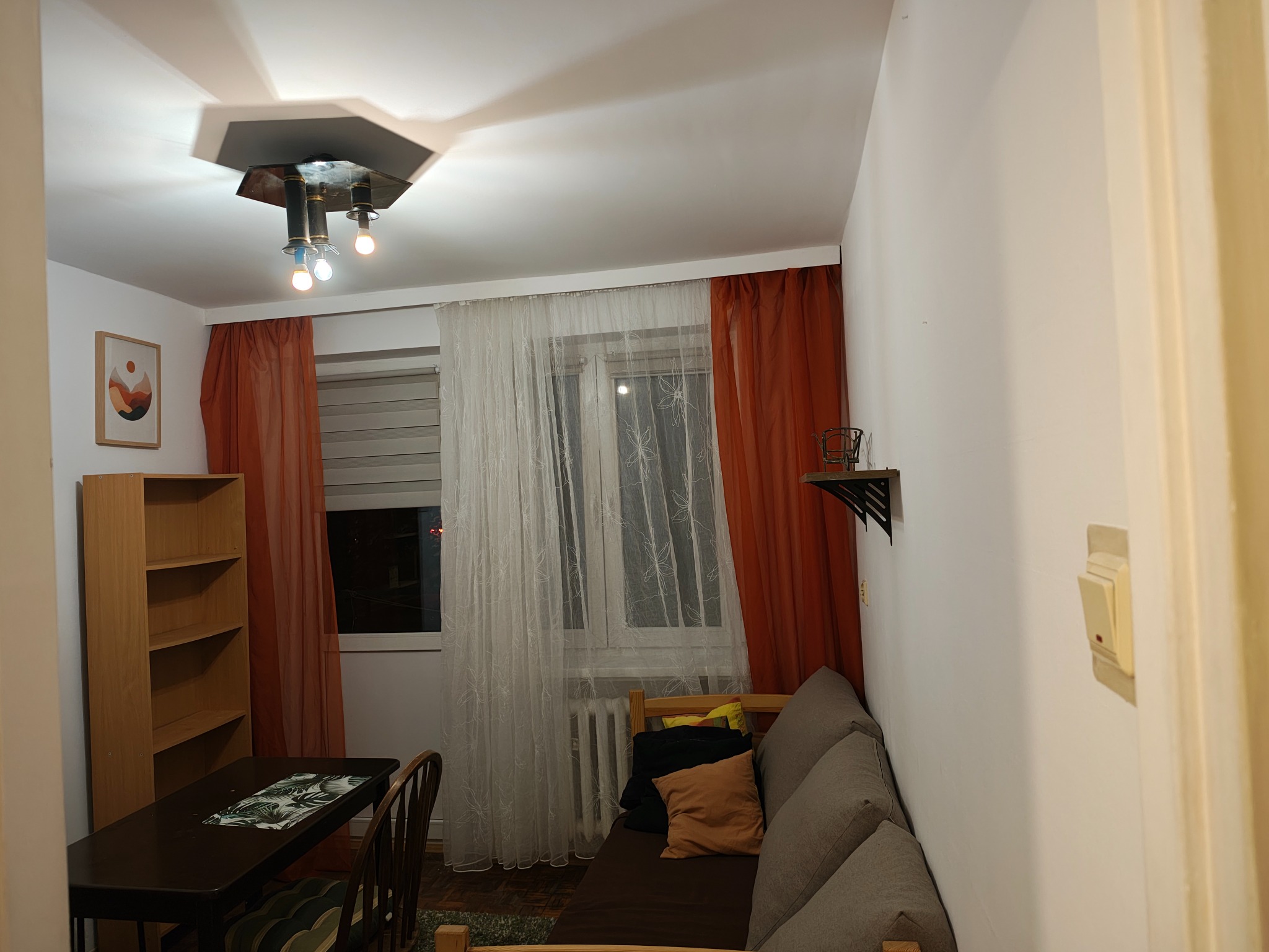 Przytulne wnętrze pokoju z drewnianym regałem, biurkiem i sofą, okno z zasłonami i designerska lampa sufitowa. Widok na ścianę z włącznikiem światła.