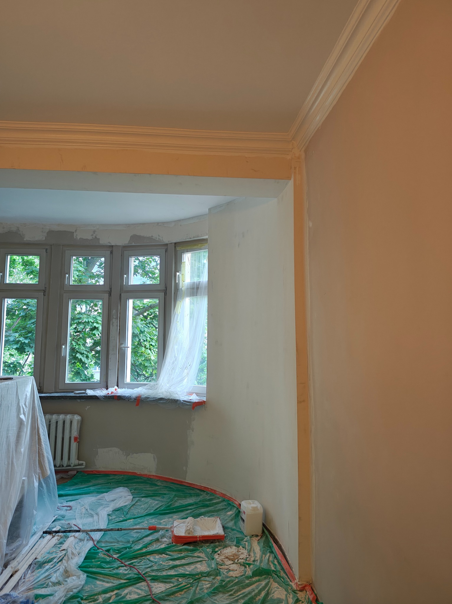Pokój w trakcie odświeżania: zabezpieczone okno z widokiem na zieleń, ściany częściowo pomalowane, podłoga osłonięta folią, widoczne narzędzia malarskie.