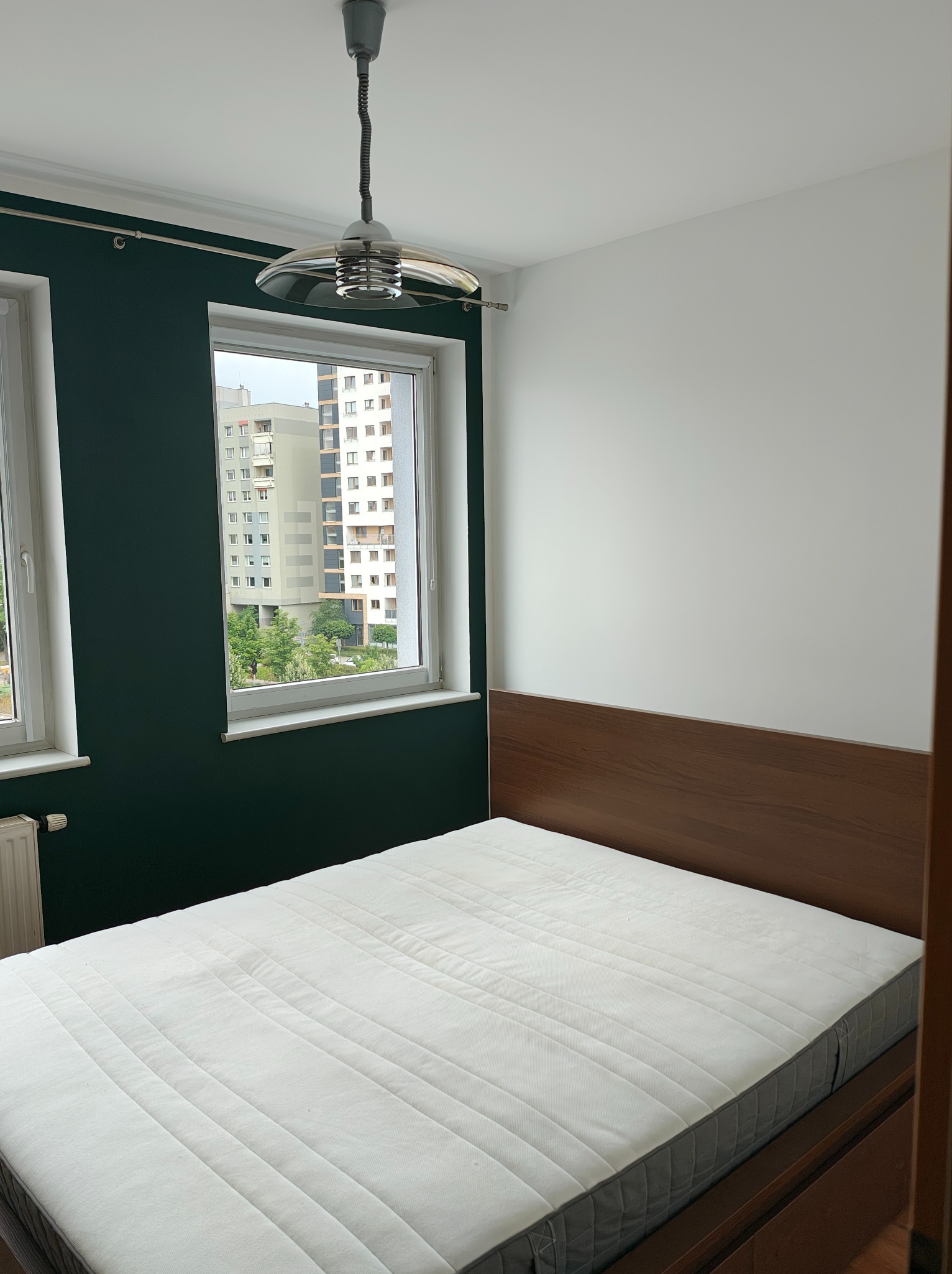 Sypialnia po malowaniu we Wrocławiu: jedna ściana biała, druga butelkowa zieleń. Widok z okna na blok mieszkalny. Nowoczesna lampa sufitowa.