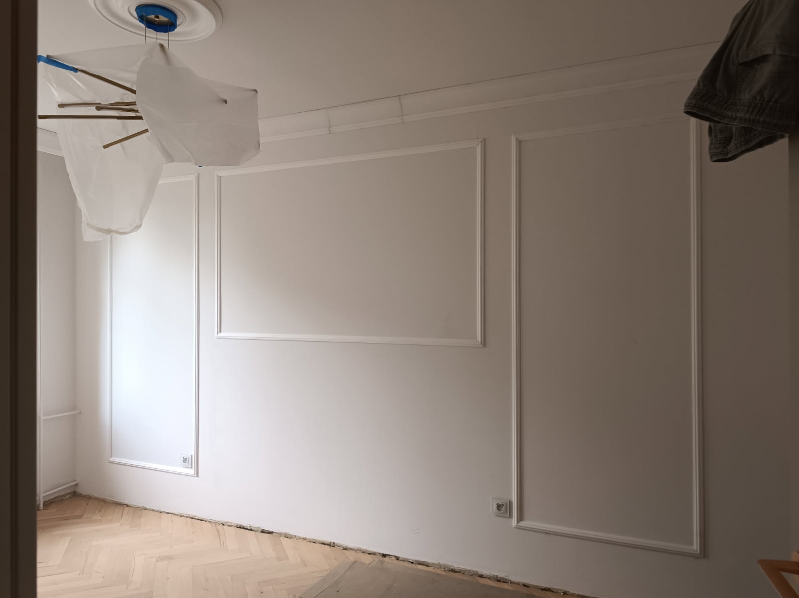 Białe ściany z dekoracyjną sztukaterią w formie prostokątnych paneli, zabezpieczona lampa wisząca, parkiet w jodełkę. Wnętrze w trakcie prac wykończeniowych.