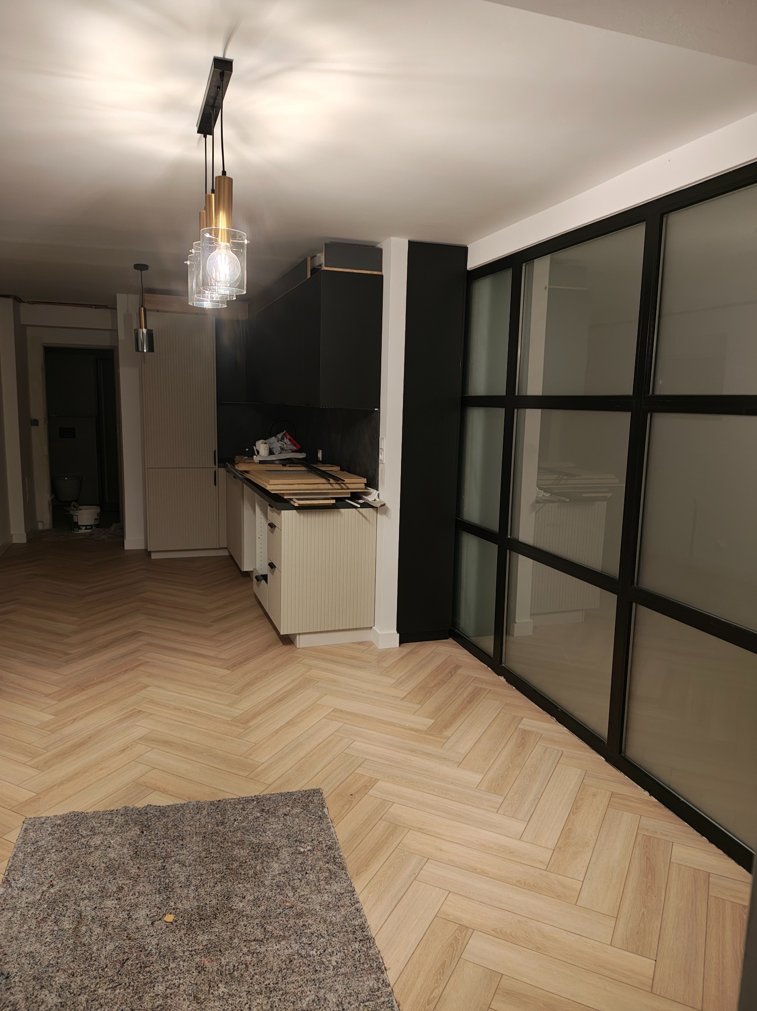 Wnętrze po remoncie we Wrocławiu: parkiet w jodełkę, czarno-biała zabudowa kuchenna, przesuwne drzwi z czarnymi szprosami i nowoczesna lampa wisząca.