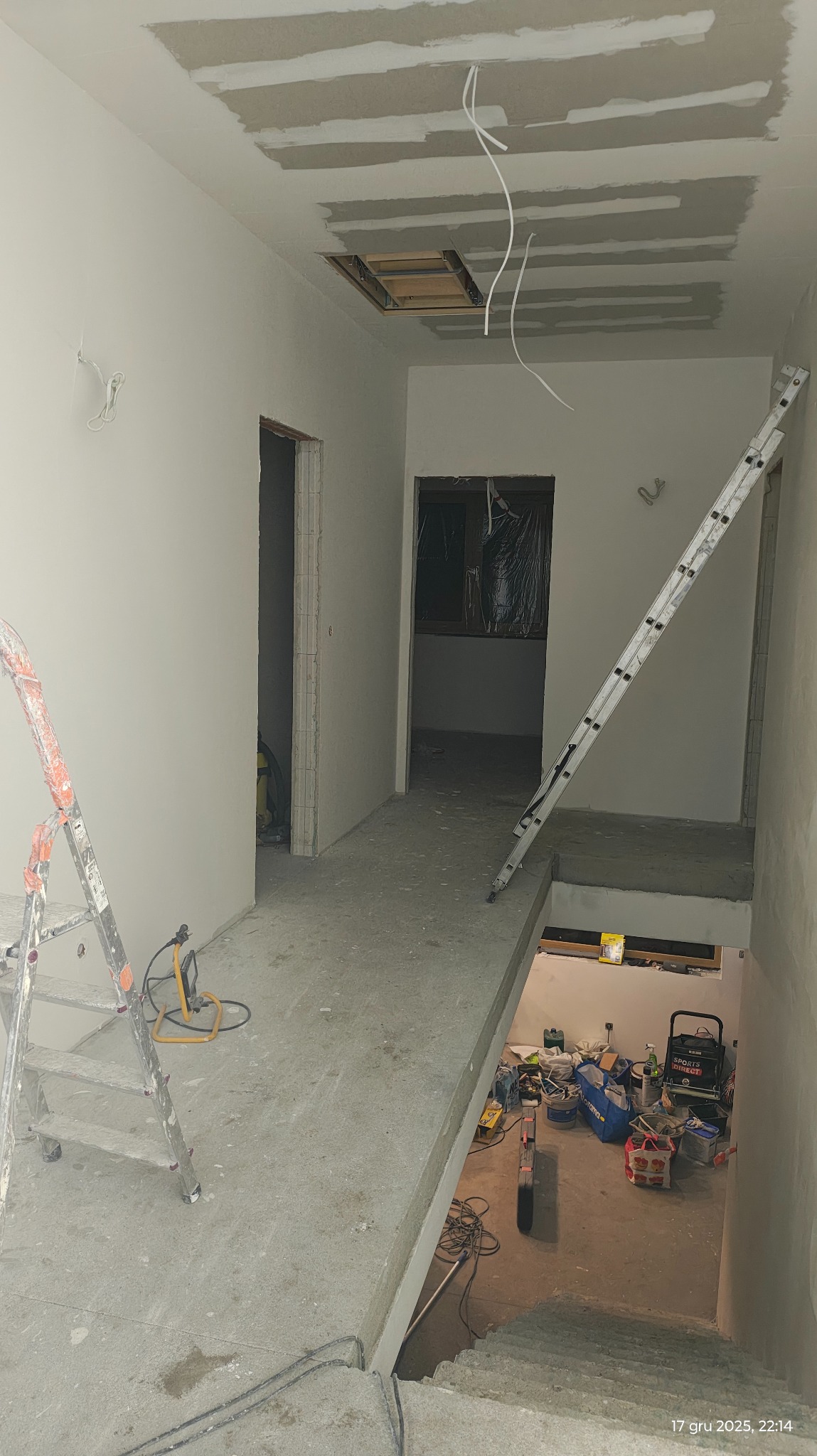 Wnętrze w trakcie prac wykończeniowych: ściany po gładziowaniu, widoczne drabiny, narzędzia i kable. Otwór w suficie z widoczną konstrukcją. Surowy stan, przygotowanie do malowania.