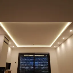 Nowoczesny sufit podwieszany z oświetleniem LED w kształcie prostokąta i punktowymi lampami, widok na okno z roletami zewnętrznymi i fragment ściany z wbudowanym elementem dekoracyjnym.