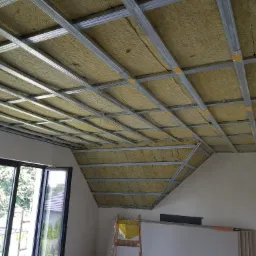 Skośne poddasze w trakcie ocieplania, widoczna konstrukcja metalowa i wełna mineralna między profilami, drabina z żółtą folią w tle, okno na zewnątrz.