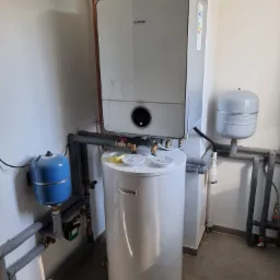 Zestaw grzewczy Bosch z zasobnikiem ciepłej wody, widoczne rury miedziane, szare rury izolowane oraz niebieski zbiornik wyrównawczy, całość zamontowana przy białej ścianie.