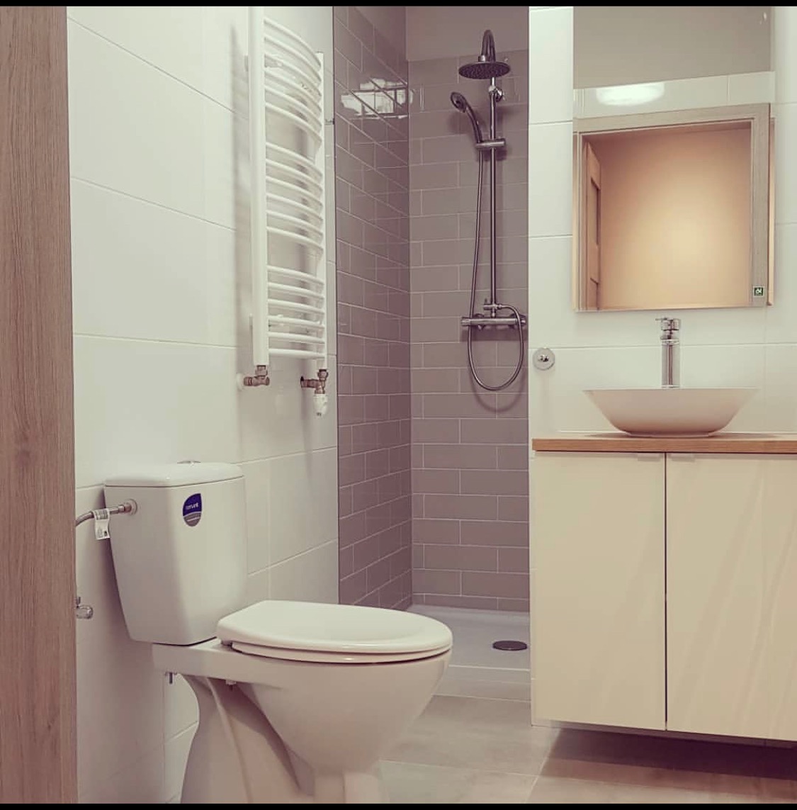 Nowoczesna łazienka z białymi i szarymi płytkami, wiszącą toaletą, grzejnikiem drabinkowym, kabiną prysznicową typu walk-in oraz umywalką nablatową na szafce z jasnego drewna.