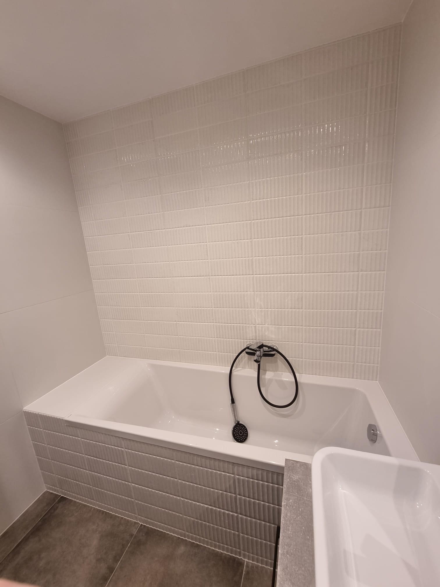 Nowoczesna łazienka z białą wanną obudowaną szarymi płytkami, białe płytki ścienne z delikatną fakturą, czarna słuchawka prysznicowa na wannie.