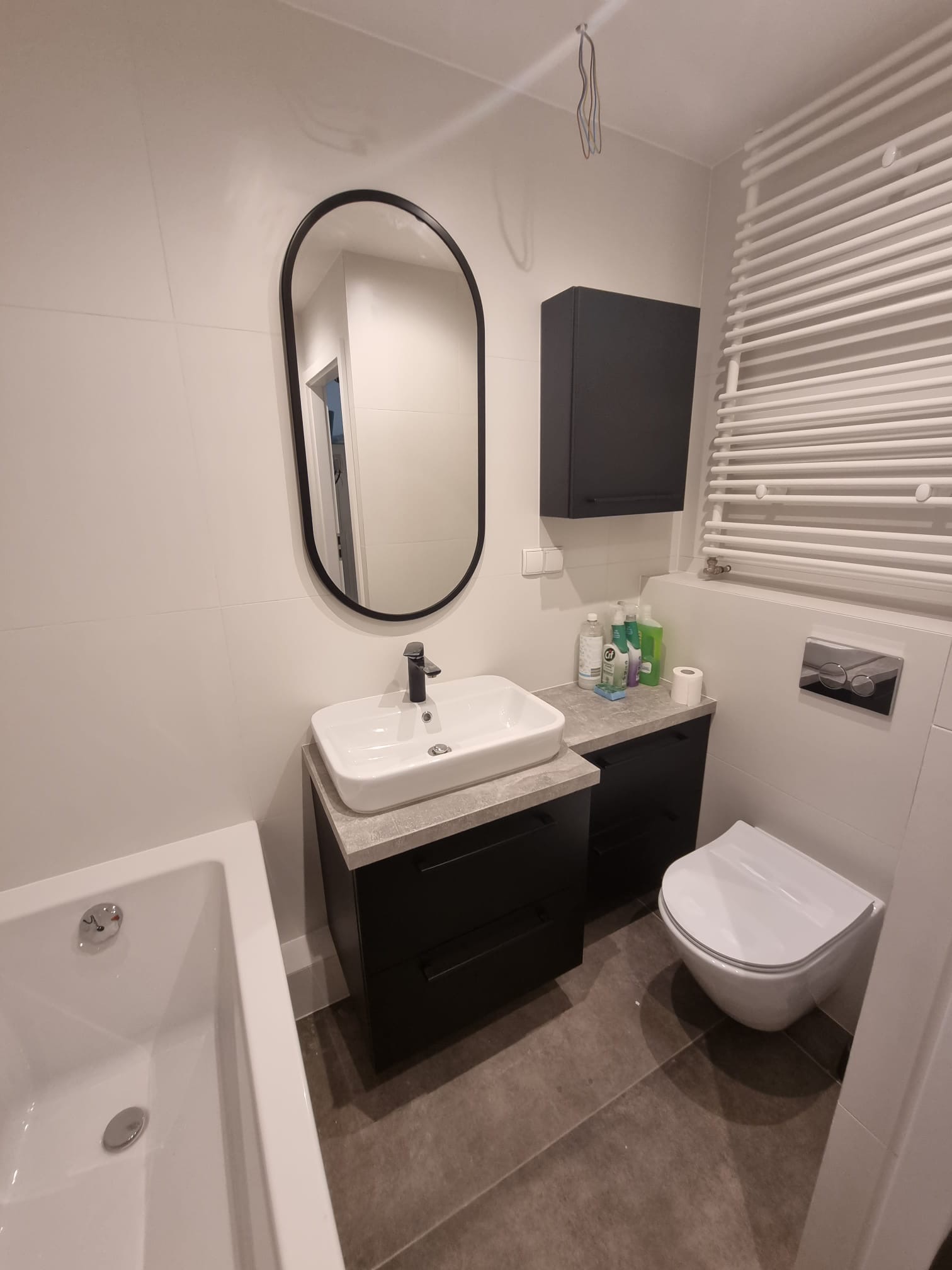 Nowoczesna łazienka z owalnym lustrem w czarnej ramie, białą umywalką na szafce z szarym blatem, wiszącą toaletą i grzejnikiem drabinkowym na białych płytkach. Szare płytki podłogowe i wisząca...