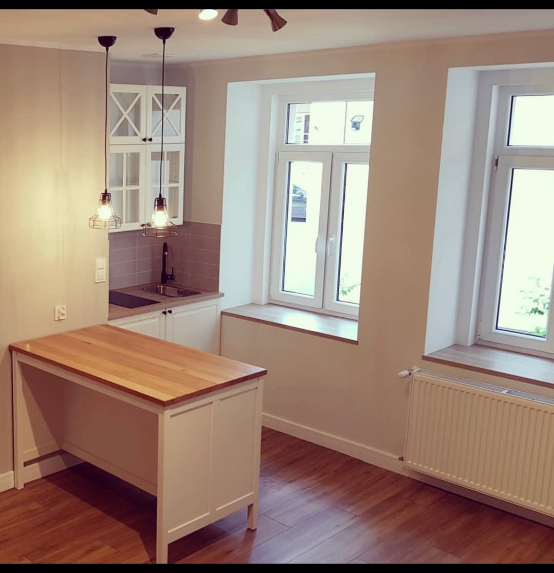 Jasna kuchnia po remoncie z drewnianym blatem, białymi szafkami i dwoma oknami z drewnianymi parapetami, oświetlona wiszącymi lampami w stylu industrialnym.